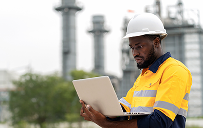 man with hard hat holding a laptop while watching the screen with a plant in background